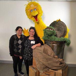 Rennata and Georgina Lopez beside the Sesame Street characters, Big Bird and Oscar the Grouch. They are posing and smiling for the camera.