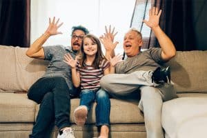 A father, daughter and grandfather sitting on a beige couch with their arms outstretched and palms out. They are pretending to be scared.