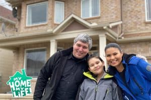 A family, mother, father, and a daughter, all smiling outside a house. They have North American Indian heritage.