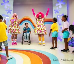 Fay cheering, arms oustretched, Fluffy cheering, beside a boy in a wheelchair, surrounded by children.