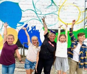 A woman (Mique Michelle) and 4 kids standing in front of a colourful unfinished mural. They are cheering.