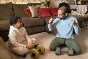 A man and his son sitting on the carpet, in front of the couch in the living room. They each have a hand barbel and are discussing how to lift them. This is a scene in an episode of "My Home, My Life".